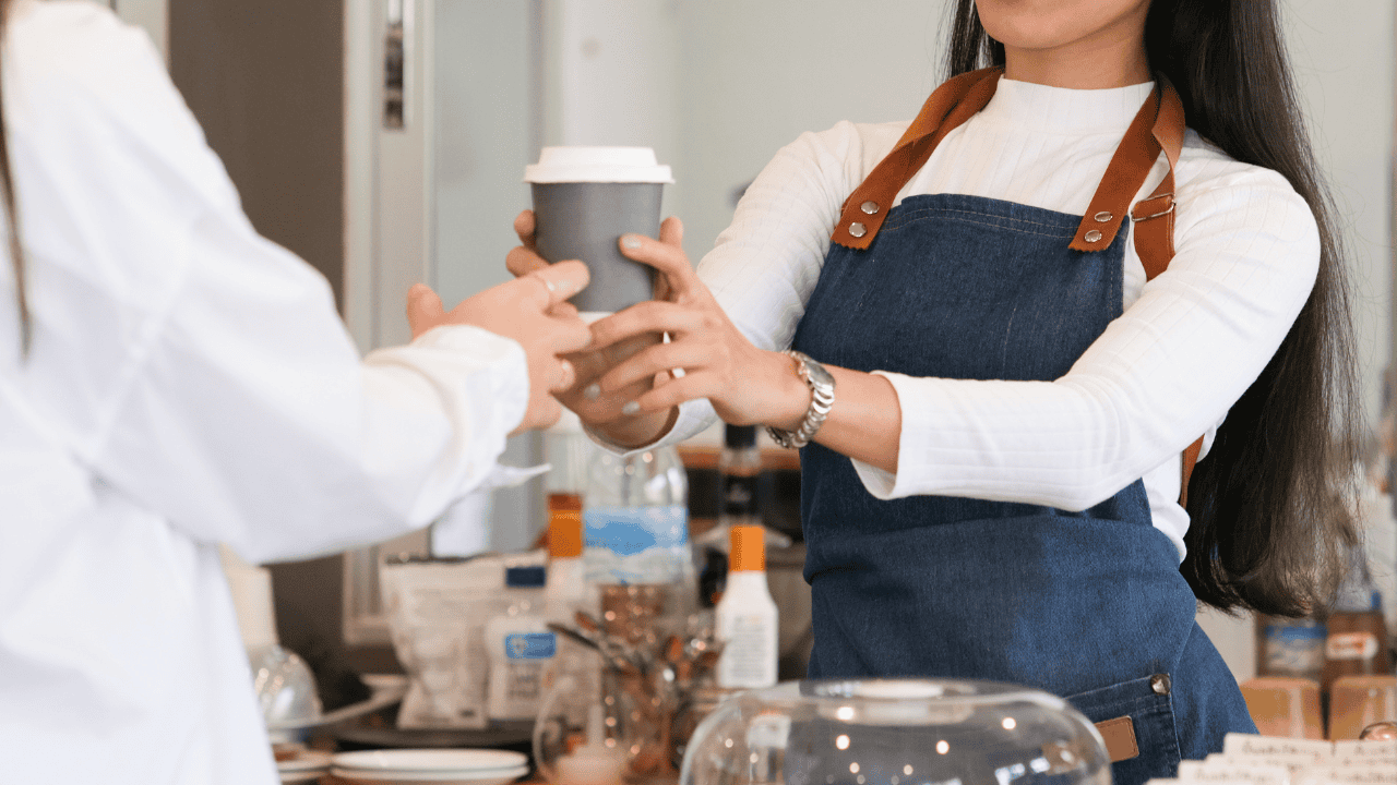 A woman in a white shirt and denim apron handing a coffee to another person in a casual coffee shop setting.