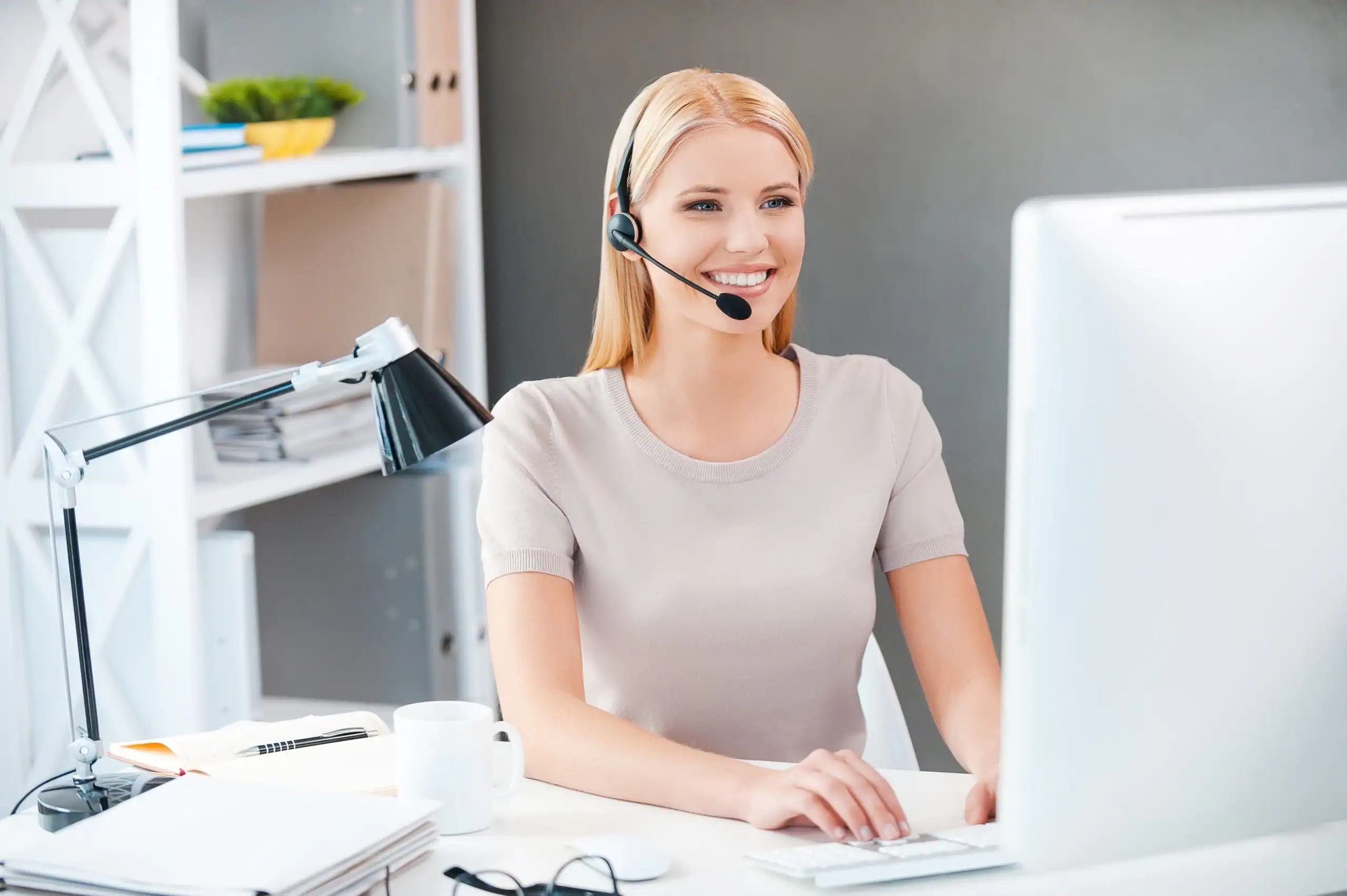Friendly woman customer support agent with headset working at computer in an office setting.